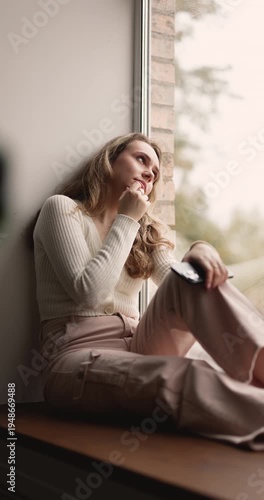 Pensive young adult woman sitting on windowsill alone at home, holding smartphone, waiting for call, feeling sadness, loneliness or boredom, thinking on personal issues during weekend leisure time