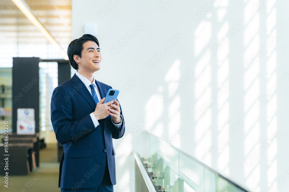 Fototapeta premium オフィスビルのロビーでスマホを持って遠くを眺める男性ビジネスパーソン