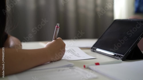closeup hand of child learn from home write on paper or young kid girl student writing doing homework with computer tablet on desk by study online class or people back to school or business learning