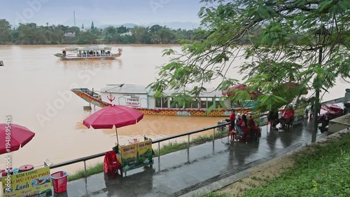 HUE, VIETNAM – 22 November 2025: People sit along the Perfume River in Hue, Vietnam, as tourist boats pass by on the water on a cloudy day.
