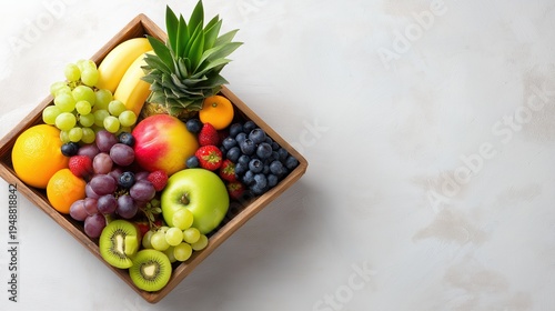Top view of wooden crate filled with variety of fresh tropical fruits including pineapple, kiwi, grapes, berries, and apples on white textured background for healthy delivery service.