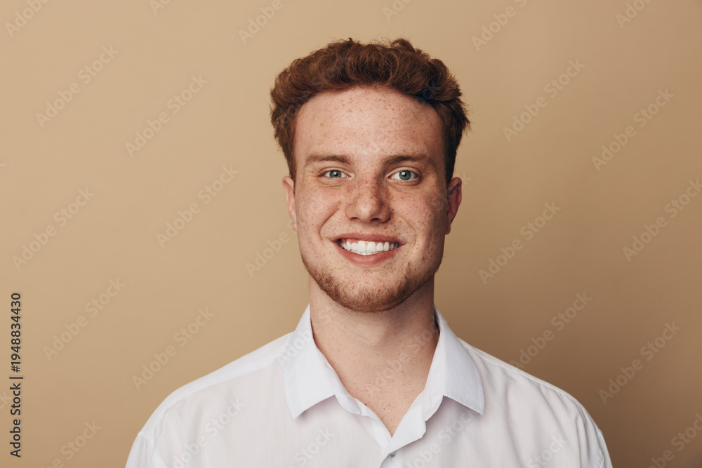 Fototapeta premium Smiling young man with curly red hair and freckles wears white shirt against beige background. Portrait of confident male model with natural skin and friendly expression in studio lighting.