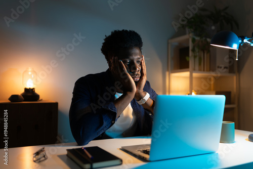 Exhausted young Black man working from home at his desk on a laptop late at night