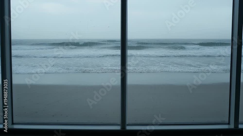 Wide beach and ocean waves under an overcast sky, framed by a window, a serene view