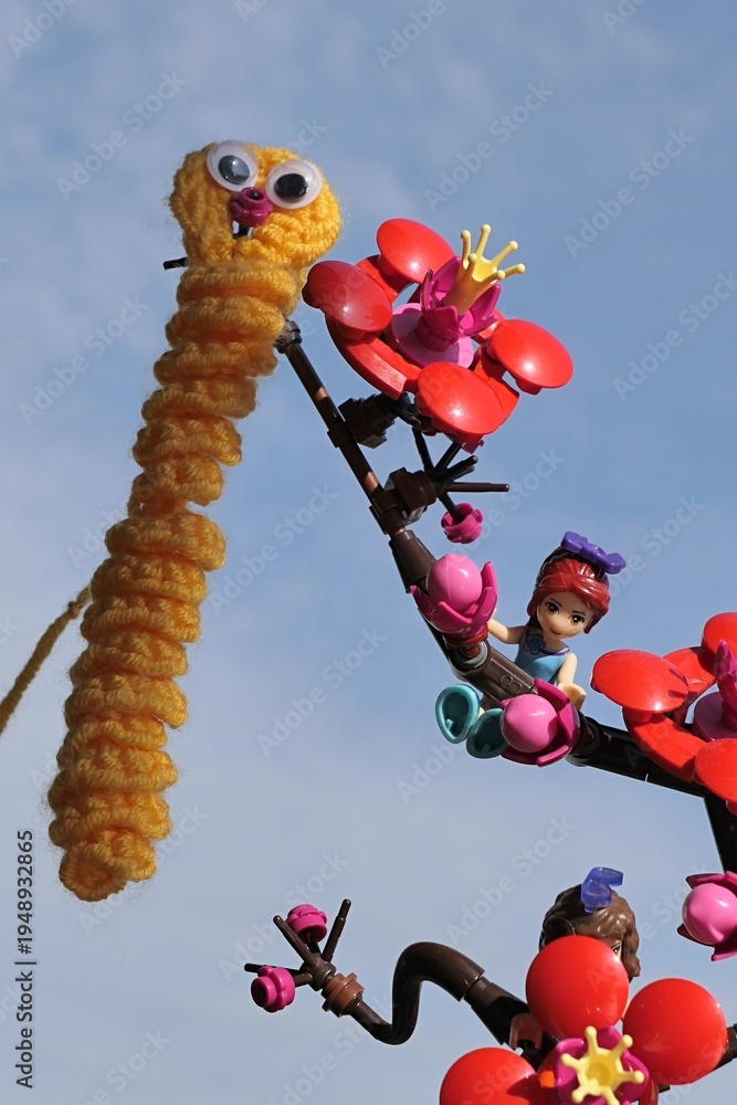 Fototapeta premium LEGO Friends figure of Mia hanging on LEGO Botanicals Plum Blossom branch, handmade knitted funny yellow caterpillar hanged on branch tip. Blue skies in background, sunlit by spring morning sunshine.