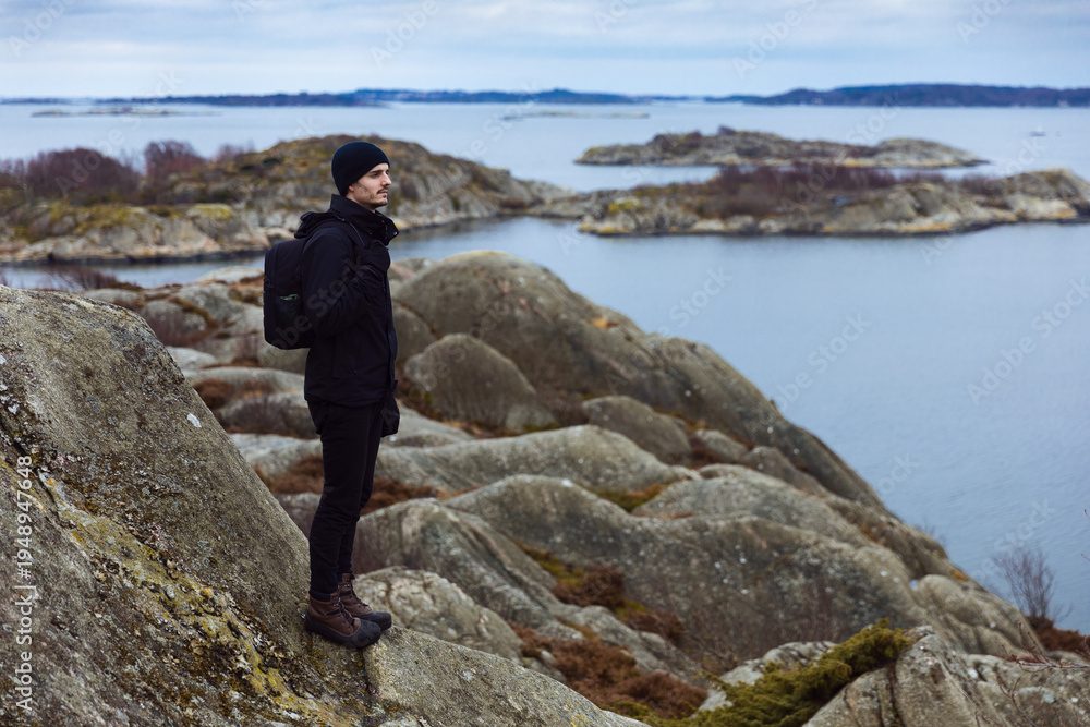 Obraz premium A man in black outdoor clothing with a backpack stands on a rocky cliff, looking out over a calm sea and scenic archipelago islands under a cloudy sky. Perfect for travel and nature themes.