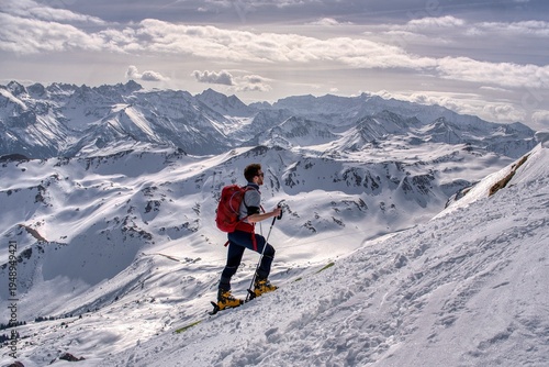 Skier Traversing Vast Snowy Alpine Slope, Lone Backcountry Guide Skiing Across Windswept Plateau Toward Jagged Peaks, Crisp Blue Sky, Sharp Shadows, Visible Tracks, Highaltitude.