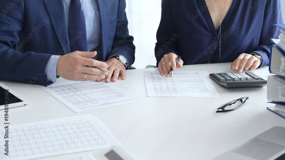 Fototapeta premium Two accountants examining financial documents using a calculator, taking notes, and discussing results, surrounded by documents and technology in a busy office environment. Audit and taxes in business