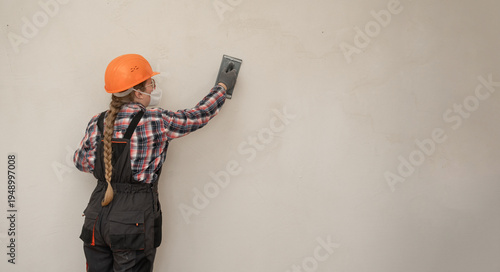 Builder woman in hardhat and protective mask sanding and smoothing walls after plastering for leveling getting it ready for painting during home renovation and construction work. banner.