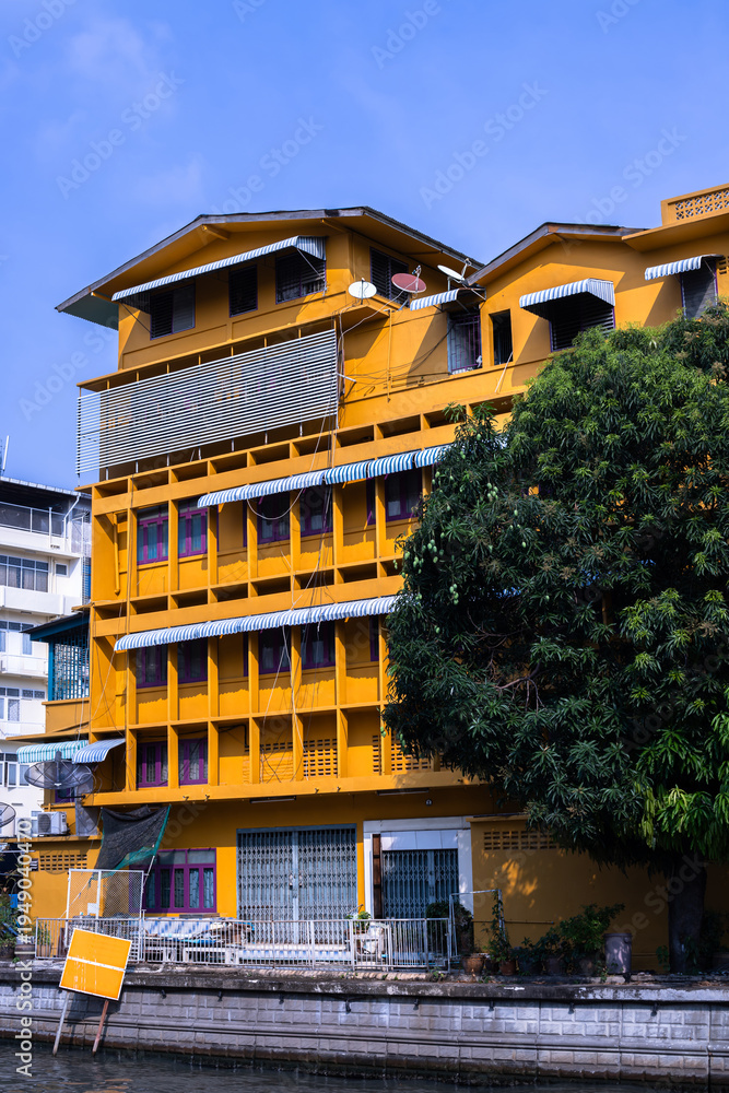 Obraz premium Yellow residential building in Bangkok near canal with tropical tree and blue sky urban architecture background