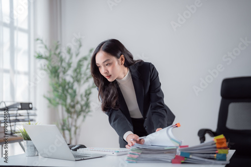 Billede på lærred Young Asian businesswoman standing at a tidy desk, organizing a large stack of d