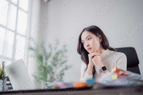 Photography Serious Asian woman sitting at her desk with laptop and papers, appearing deep i