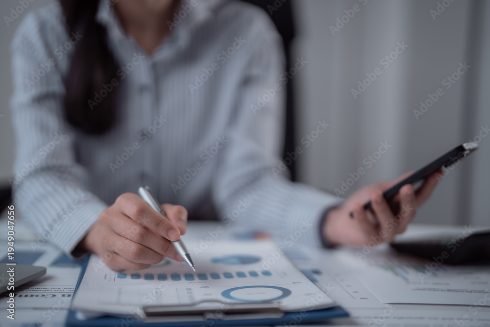 Fototapeta premium Businesswoman's hands reviewing financial report graphs and charts on a clipboard, using a pen to indicate figures while holding a smartphone for additional data