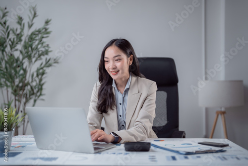 Young Asian businesswoman sitting at her desk, typing on a laptop, and looking happily at the screen while managing work in a corporate office environment