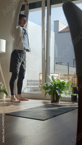 Man standing barefoot and stretching arms near large window in sunny living room with exercise mat and plants. Concept of home workout warmup wellness routine and healthy lifestyle.