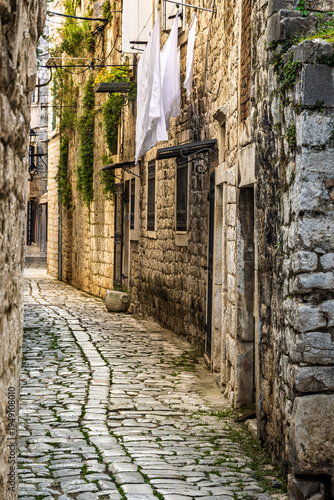 Narrow stone alley with cobblestone street and hanging laundry in historic old town Trogir, Croatia, authentic Mediterranean architecture and traditional Dalmatian atmosphere.