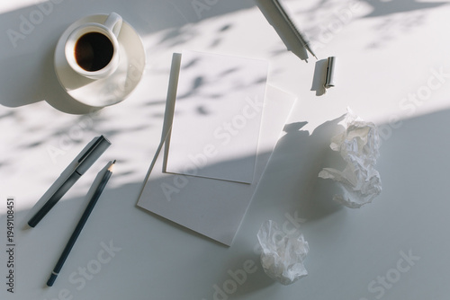 Overhead view of blank paper mockups and a cup of coffee with crumpled paper bits symbolizing a creative brainstorming process under leaf shadows.
