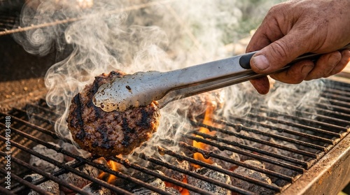 Fotografie Close up of tongs flipping a juicy smoking burger patty on a hot outdoor barbecue grill