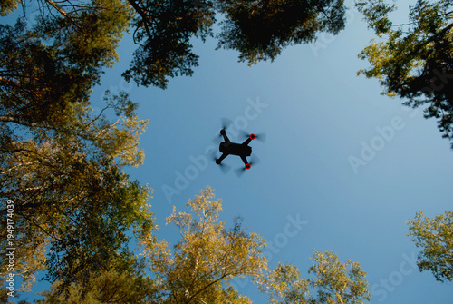A small drone in flight captured from below. The dark silhouette of the quadcopter contrasts with the bright blue sky, with red navigation lights visible on its rear. Treetops frame the scene