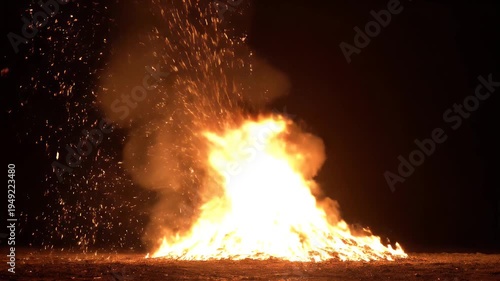 A large, bright bonfire burns in the dark night, sparks and embers flying upwards