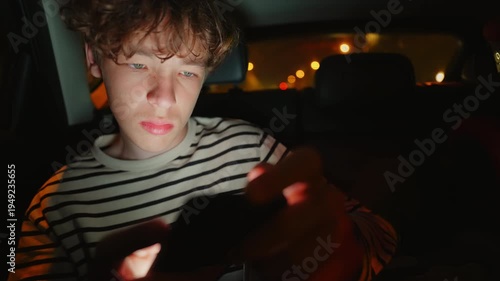 A young person sits in a car at night, illuminated by the glow of a mobile phone screen. They intensely use the device, eventually showing signs of discomfort or fatigue