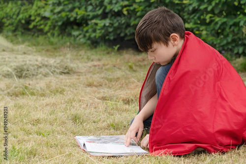 A boy is reading a book intently. He is sitting on the grass, wrapped in a red sleeping bag.