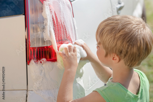 A boy is washing the taillights of a car. He rubs it sponge wiht foam.