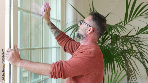 Middle-aged man with beard and glasses cleaning a glass door with a pink cloth in a bright modern home interior, representing household chores and everyday domestic life