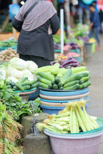 A bustling local market scene showcasing piles of fresh cucumbers, corn, and leafy greens being sold by a vendor