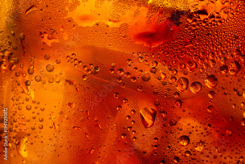 A close-up image of condensation droplets on a glass of refreshing amber-colored, cold cola drink,Macro shot of vibrant orange and red liquid texture behind a glass surface covered in fresh condensati