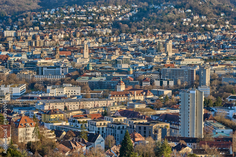 Naklejka premium Panoramic view of Stuttgart city center in Germany during spring with warm golden evening light. Dense urban architecture, hills and residential districts captured from an elevated viewpoint.