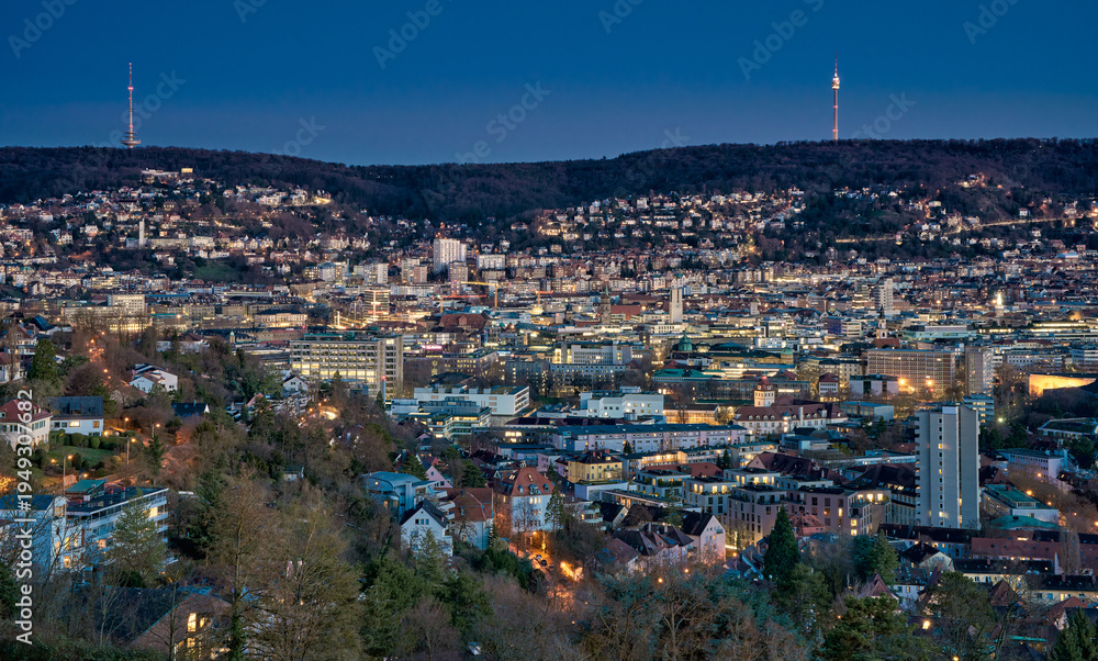 Fototapeta premium Panoramic blue hour view of Stuttgart city center in Germany with glowing city lights, dense urban architecture and residential hills. Television towers visible on the surrounding hills.