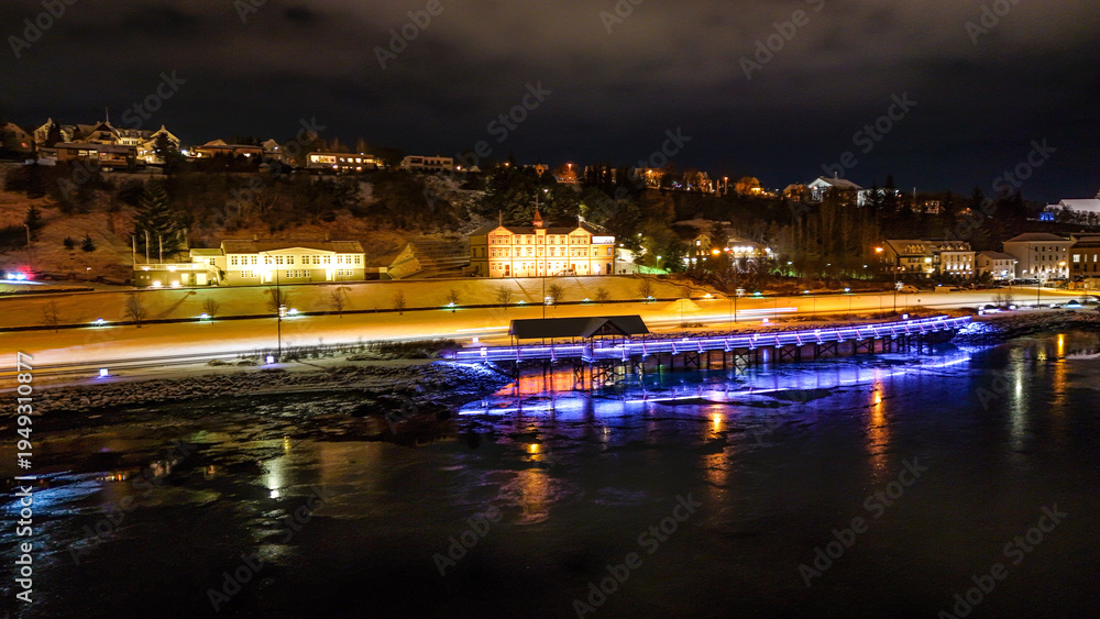 Fototapeta premium Drone view of Akureyri in North Iceland at night with illuminated bridge and city lights reflecting on water. Beautiful aerial scene of Akureyri harbor showing urban landscape and coastline in Iceland
