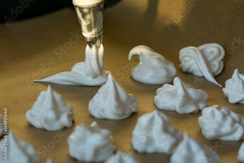 Close up of a pastry bag piping small meringue drops onto parchment paper on a baking tray.