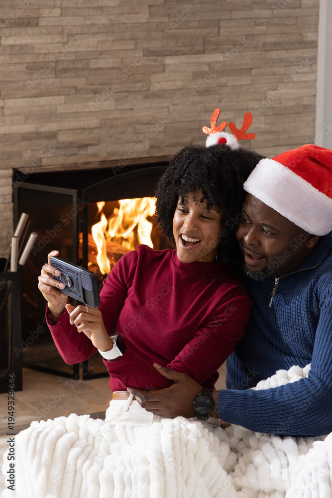 Fototapeta premium African American couple sitting by fireplace, taking selfie on phone, Santa hat, snuggling blanket