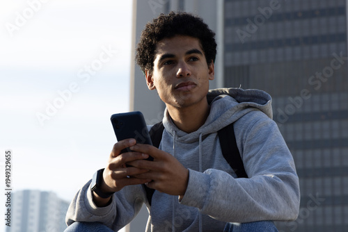 Tableau sur toile African American teen male holding smartphone and gazing right on roof wearing h