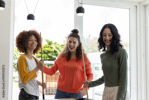 Diverse female friends playing pool at home, holding cues around green felt table near windows