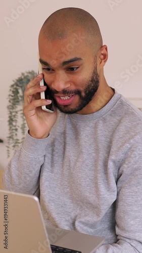 Vertical HD video. Young adult African American man telecommuting from home office using laptop. Remote worker focused on digital tasks, having a phone call in modern workspace. Business concept