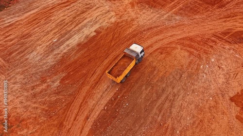 Truck loaded by the red soil goes slowly by the rocky area. Arid mountains with poor vegetation in the backdrop. Aerial view.