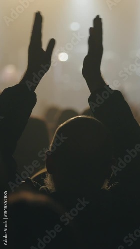 Rear view of an audience member silhouetted against stage lights, raising and clapping hands at a concert, worship service, or public event. Emotional atmosphere with changing light - vertical footage