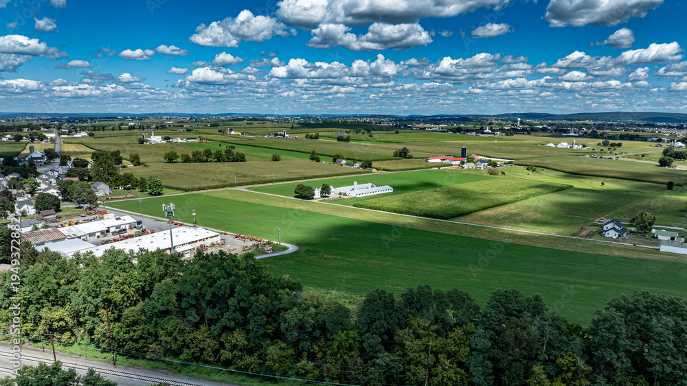 Fototapeta premium The scene shows a broad view of fields and farms located near a small town. Green fields stretch across the landscape under a bright blue sky with scattered clouds. Buildings and trees are visible.