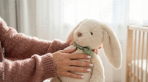 Adult woman in cozy sweater holds a soft plush bunny toy with a green bow, sitting in a well-lit room with a crib and sheer curtains in the background