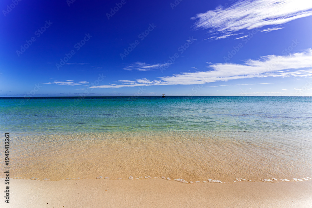 Obraz premium Pristine sandy beach with crystal-clear turquoise water and gentle waves under a deep blue sky in the Florida Keys, featuring a distant boat on the horizon and a serene tropical atmosphere