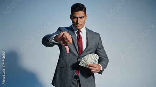 Businessman in sharp gray suit expressing strong disapproval with a thumbs down gesture while holding a significant fan of United States hundred dollar bills cash.