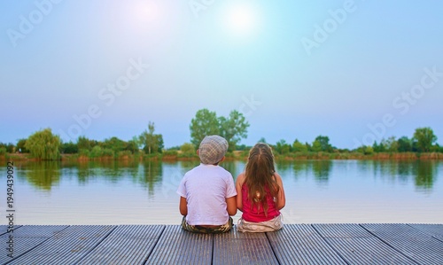 Fotografia A little boy wearing a flat cap and a cute little girl are watching the sky on a wood pier on the shore of a lake