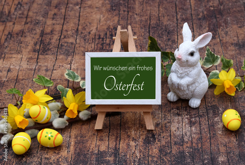 Kleiner Staffelei mit grünem Schild und weißem Rahmen mit der Aufschrift 'Wir wünschen ein frohes Osterfest' neben einer weißen Hasenfigur, gelben Narzissen und bemalten Ostereiern auf Holzuntergrund.