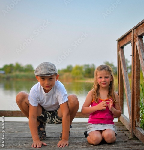 A cute small boy with a flat cap and a sweet little girl are sitting on a wooden pier on the shore of a lake. Small children are sitting on a pier at the edge of a pond. 