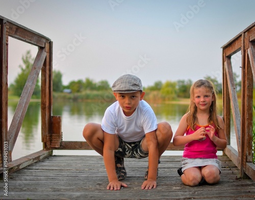 A cute small boy with a flat cap and a sweet little girl are sitting on a wooden pier on the shore of a lake. Small kids are sitting on a pier at the edge of a pond. The concept of a childhood.