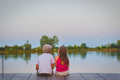 A little boy wearing a flat cap and a cute little girl sit with their backs turned on a wooden pier on the shore of a lake. Small children sit on a pier at the edge of a pond. The concept of a childho