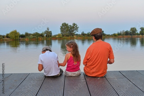 Children sitting on pier. Three children of different age - teenager boy, elementary age boy and preschool girl sitting on a wooden pier. Summer and childhood concept. Children on bench at the lake. 